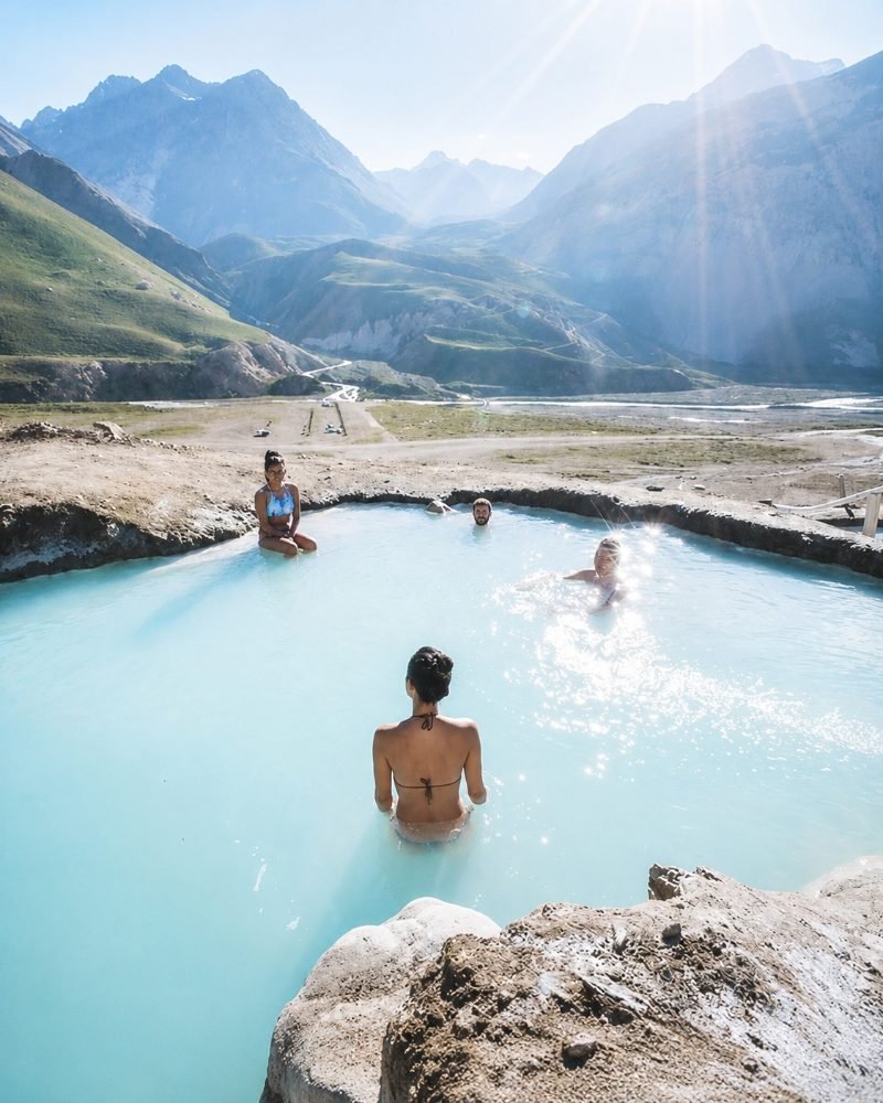 Group of friends soaking in turquoise thermal pool with dramatic Andes sunburst above
